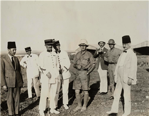 A group of men stand in the desert. Some are wearing white suits with Turkish hats. Some are wearing pilot helmets. Some holding drinking glasses. 