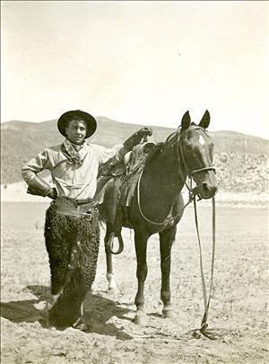 A cowboy stands by his horse near Bridgeport, his hand resting on the saddle. He is wearing a cowboy had, heavy gloves, a neckerchief, and what appear to be wool chaps.
