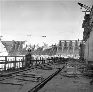 A well-dressed woman stands in the foreground at the Chief Joseph Dam jobsite. In the background looms the huge dam, still under construction when photo was taken in 1955.