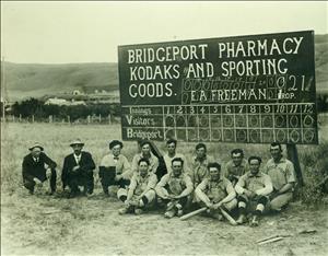 A group picture of Bridgeport baseball team in about 1912, with nine men in uniforms and the others in street clothes. They are sitting in front of a scoreboard sponsored by the Bridgeport Pharmacy that indicates a recent completion of a 21-inning game 