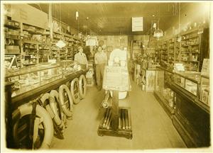 Interior photograph of Bouska and Son hardware store in Bridgeport, with shelves crowded with a large variety of good and, leaning against the counter to the left a number of automobile tires. Standing in the middle are two men, presumably Mr. Bouska and his son.
