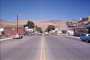 Color image taken in 1976 of the view looking north-west from Columbia Avenue and 12th Street in Bridgeport, Douglas County, WA. There are several businesses on either side of the street. On the far right is the Bridgeport IGA Supermarket. On the left, the second building is the Douglas Federal Credit Union. Automobiles are parked on either side of the street. Dry barren hills are visible in the distance.