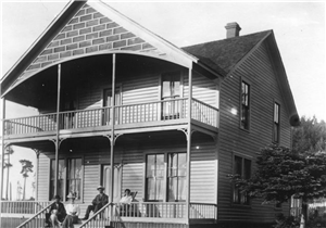 Large two-story frame house with four people and a dog sitting on front porch and steps leading up to it