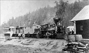 A group of men stand in front of a steam locomotive. Behind the train a hill covered in trees.