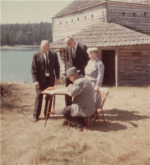 Older man signing papers at a table set up in a field in front of an old blockhouse and view across a bay; two men in suits and an older woman standing behind table watching