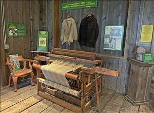 Wooden loom in room with floor and walls of unfinished wood; jackets and signs hang on wall behind the loom