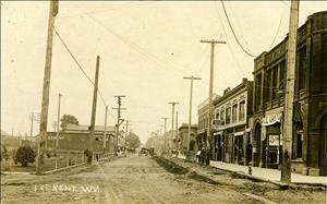 Photo taken looking down 1st St. in Kent, King County, WA, ca 1908. The street is unpaved. There are a few wagons on the street. On the right side is the Kent Cafe, Fashion Hotel, Kent News and Book store. Telephone poles run the length of the street on both sides. People are on the sidewalk shopping. A fire hydrant and barberpole are visible.