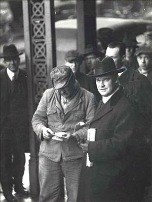 Washington Governor Ernest Lister speaking with unidentified men as others look on. Lister is looking holding a folded newspaper and looking at the camera, while the man to whom he is speaking appears to be reading something. The photo is date January 11, 1919, which was the year of Lister's death.