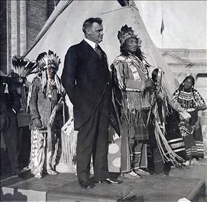 Governor Ernest Lister, in suit, stands on podium with Chief Many Tail Feathers of the Blackfoot Tribe, wearing what appears to be a crown and holding a staff. To the left stand two Native Americans in full-feathered headgear, and to the right is an Native-American woman in traditional garb. A teepee forms the backdrop of the photo. Governor Lister was being "adopted" into the Blackfoot Tribe by the chief. The photograph was taken at the Panama-Pacific International Exposition in San Francisco, 1915
