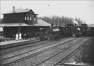 Depot with two steam engines on railroad tracks and group of men posing on platform 