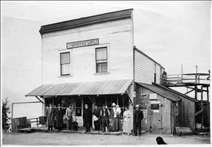 Small frame building with sign reading "US Post Office Lopez" above 2nd-floor window and line of people standing under front awning