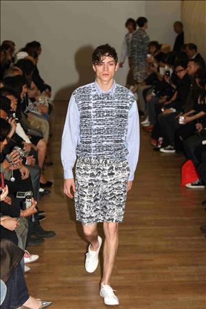 A gaunt man wearing a shirt and shorts covered in black sketches walks down a fashion runway between crowds of observers with cameras 