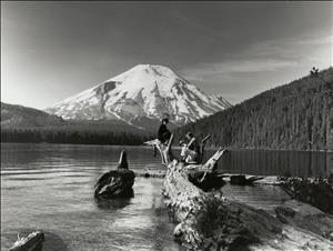 Black and white photo of a man and a woman sitting on a log on a lakeshore with a snow capped mountain and forest in the background