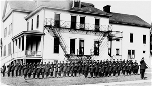 Large group of boys and young men all wearing military uniforms hold their right hands to their heads in salute in front of a large wooden building