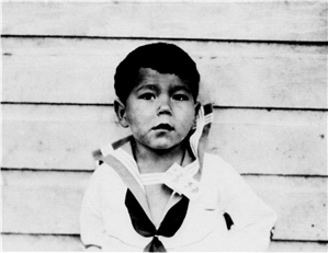 A Native American child in a sailor suit stands in front of a wooden wall