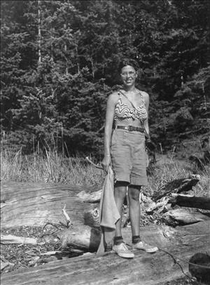 Randlett smiling in glasses, crop top and belted shorts, standing on a log in a forested area