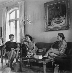 Three women sitting on a couch and chair around a coffee table beside a Palladian window and beneath a candelabra and large, framed painting during the day
