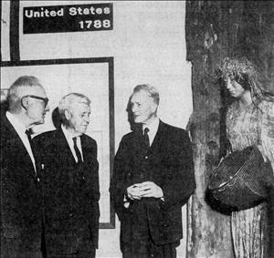 Bloedel in a suit and tie speaking with two other men in front of a museum display with the life sized mannequin of an indigenous woman carrying a woven basket, below a sign reading "United States 1788"