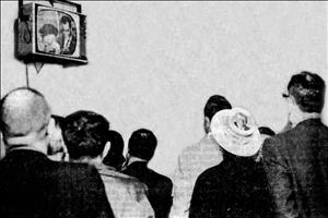Group of people, including someone wearing a straw boater with the name and picture of John F. Kennedy,  standing and watching a small television mounted high on the wall, displaying a smiling Richard Nixon