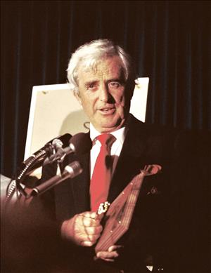 An older Ketcham holding an award, speaking in front of microphones at a podium