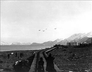 Four single propeller seaplanes fly above a range of snowcapped mountain peaks and a large bay. On the ground, people stand on the beach and on railroad tracks looking up at the planes. 