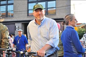 McGinn holding a bicycle outside wearing a "Seattle Cricket Club" baseball hat