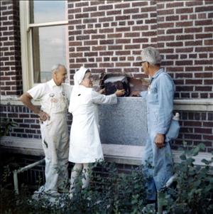 Two white men in work clothes and a white woman in a nurse's uniform hold a box next to an exterior brick wall