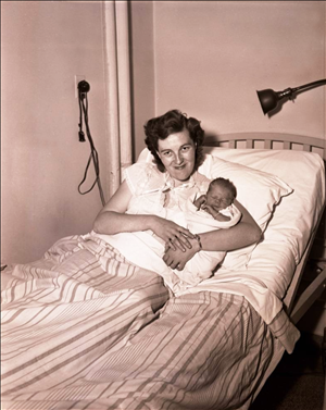 A white woman in a bed holding a newborn baby under one arm