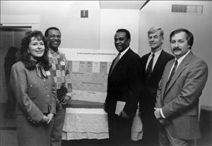 Four men and one woman in suits stand in front of an infographic smiling at the camera