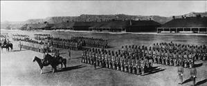 Nine rows of soldiers stand in front of barrack buildings