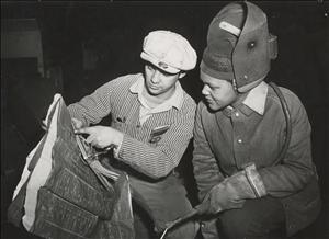 A person in a white hat shows a large sheet of paper with diagrams on it to a Black woman in a welding helmet holding a welding torch