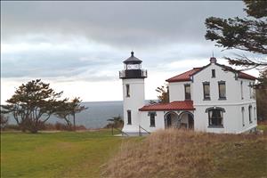 Two-story white painted building with lighthouse tower attached on the left. There are trees to the left of the lighthouse and lawn in front of it. Admiralty Inlet is in the background. 