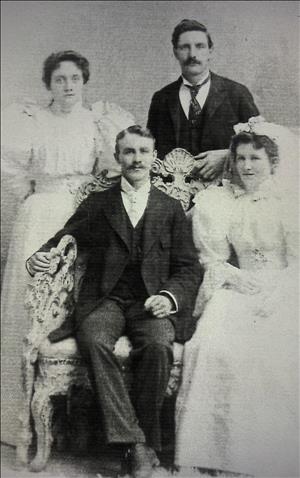 Men wearing suits and ties, women in white dresses, one couple seated in ornately carved chair, others standing behind 