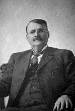 Portrait of a seated white man in a suit and tie with eyeglasses and a mustache