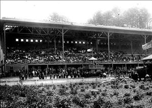 Photo of large grandstand filled with people at The Meadows racetrack in Georgetown, King County. The structure is not finished and construction debris can be seen in the foreground. Men are standing on the track and two American flags are flying