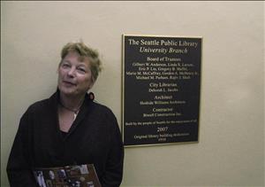A middle aged white woman smiles at the camera standing next to a large plaque on a wall. 