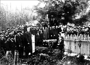 A large crowd of white and Asian men wearing suits stand in the woods next to a white fence and a coffin