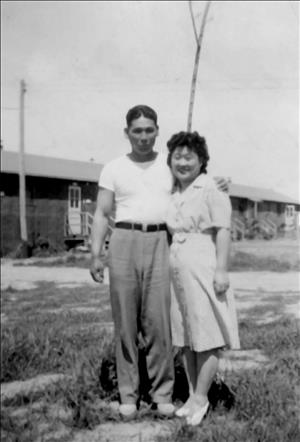 An Asian man and woman stand arm in arm in front of a one-story wooden house