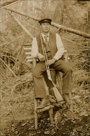 A Japanese man holds a rifle in the woods with dead waterfowl at his feet. He is wearing a hat and a suit. 