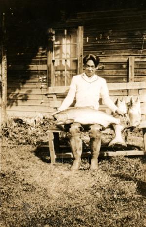 A Japanese-American young man sits on a bench holding a large fish. There are more fish on the bench. 