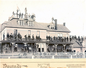 Large mansion with dozens of bicyclists in front, on the balconies, and on the roof. 