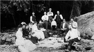 A group of men and women in suits and dresses having a picnic on a rocky beach