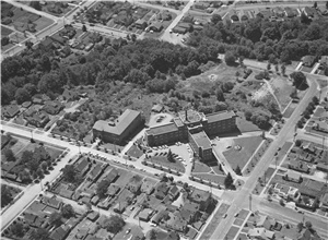Bird's eye view of a large brick building shaped like a cross surrounded by trees and houses