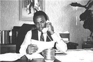 Seattle City Councilman Norm Rice sitting at desk in his office, talking on telephone while looking at a paper. Coffee cup on table, painting on wall behind him, and what appears to be a sculpture to his left