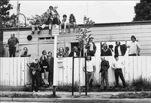 Early volunteers to KRAB-FM radio pose together outside their first studio, a former donut shop in Seattle's University District. Some stand on sidewalk, some look over a fence, and a few perch on the roof.