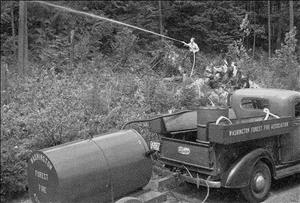 A person spraying water into forested land connected to a truck that reads Washington Forest Fire Association