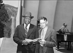 Hawley and Robeson holding two sides of the same handbill in front of a large, concrete bandshell with two men smoking and looking on in the background