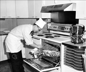 Luke in chef's whites leaning over an oven in a kitchen inspecting food wrapped in tin foil