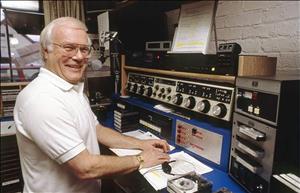 An older Bernier standing in front of a radio announcer console and microphone and smiling