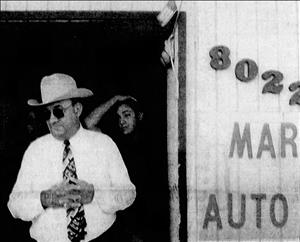 Marks in a cowboy hat, sunglasses and hands clasped together emerging from a door beside signage reading "8022, Marks, Auto"